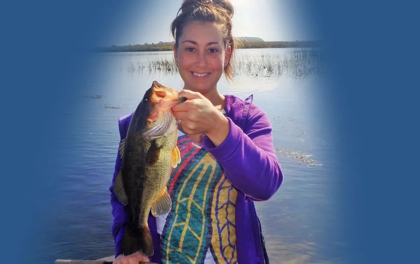 Woman smiling, holding a largemouth bass with a lake in the background.