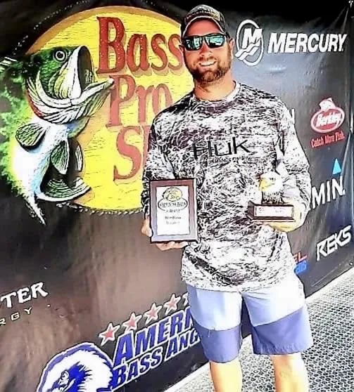 Man in sunglasses and camo shirt holding two fishing awards in front of a Bass Pro Shops banner.