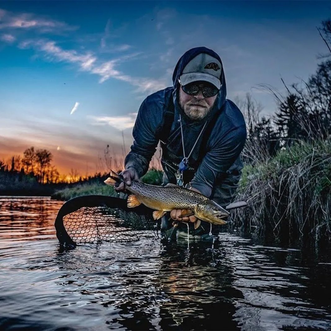 Smiling man in a dark jacket holding a large, spotted Brown Trout in a wooded stream.