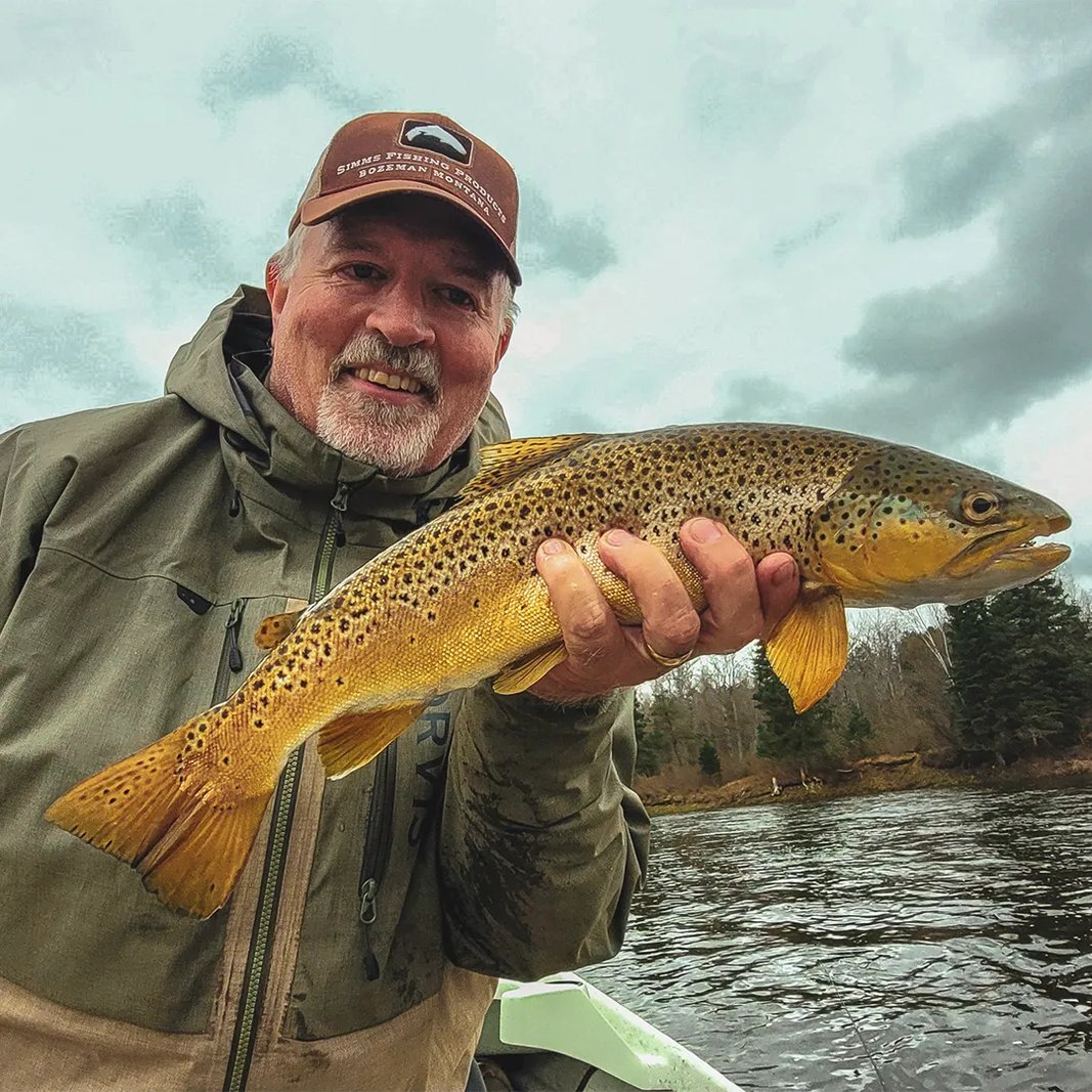 Angler in waders holding a Brown Trout in a river at sunset with an orange sky.