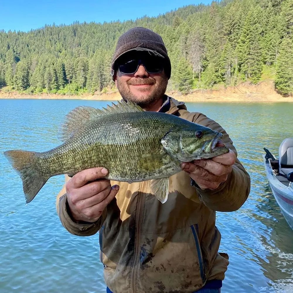 Man in camouflage holding a Smallmouth Bass while kneeling by a lake shore.