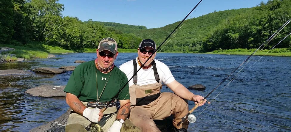 Two fly fishermen in waders sitting on rocks in a wide river.