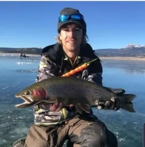 Angler kneeling on ice, holding a large rainbow trout with mountains in the distance