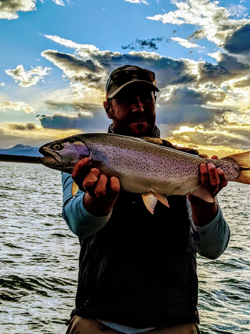 Angler holding a rainbow trout against a dramatic sunset sky over a lake