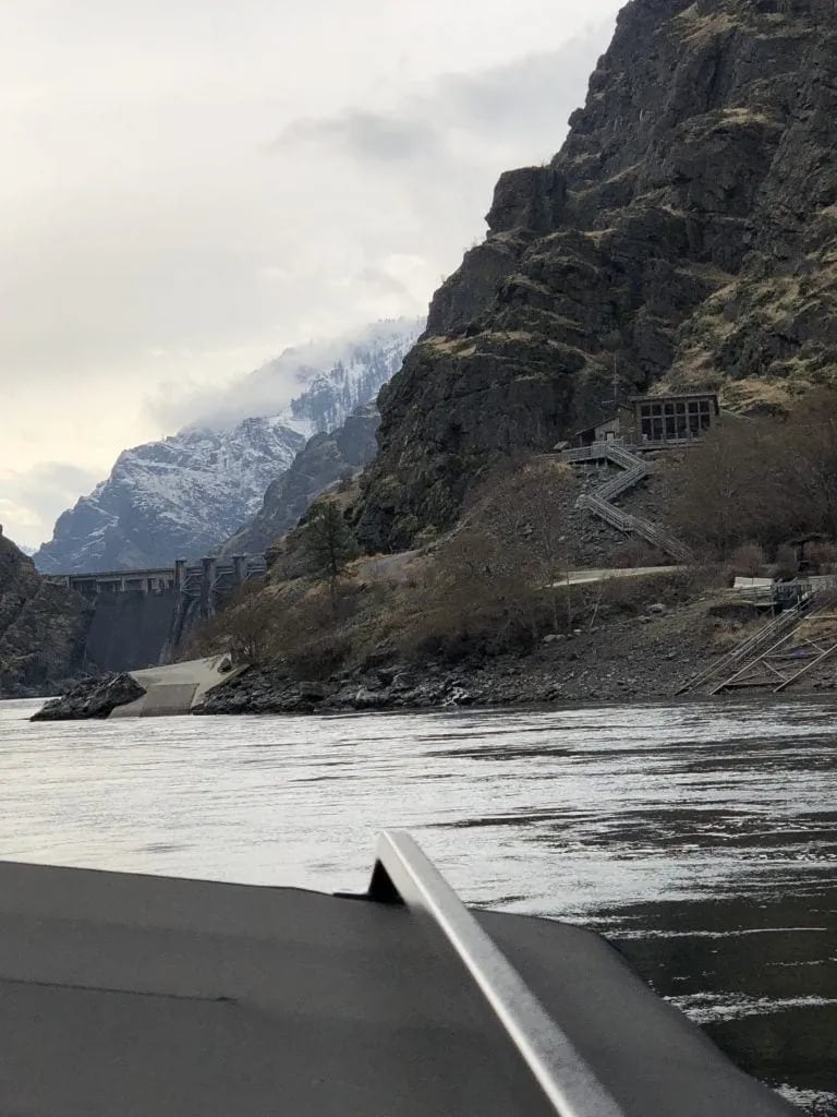 River view from a boat with steep mountains and a dam in the distance under a cloudy sky