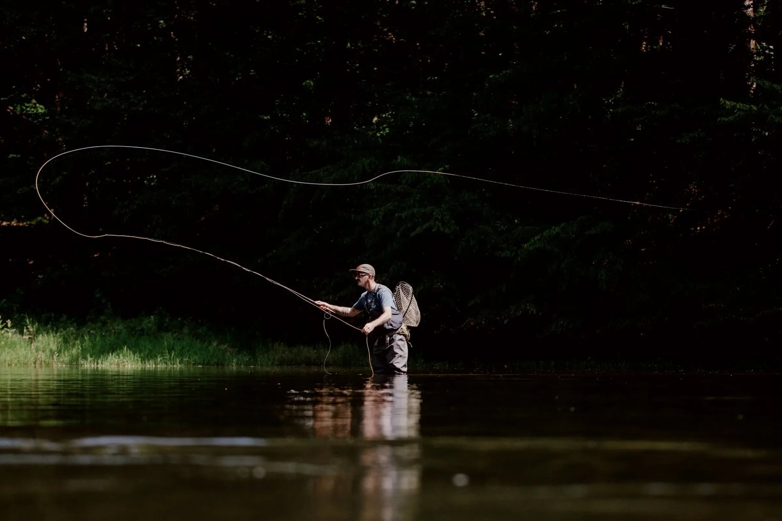 Angler in waders fly casting in a dark river with dense trees.