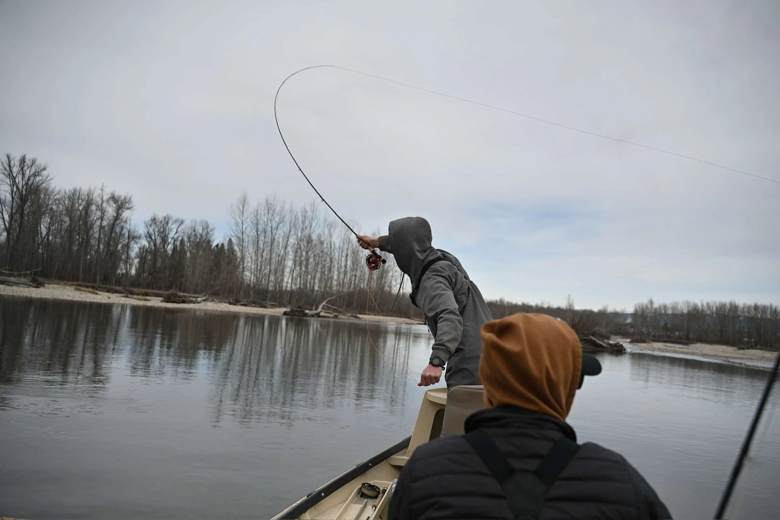 Angler casting a fly rod from the bow of a drift boat on a wide river under a cloudy sky
