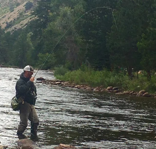 Angler wading in a river, casting a fly rod with mountains in the background