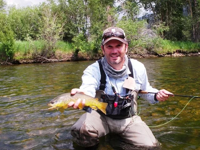 Angler in waders holding a Brown Trout in a river, smiling at the camera