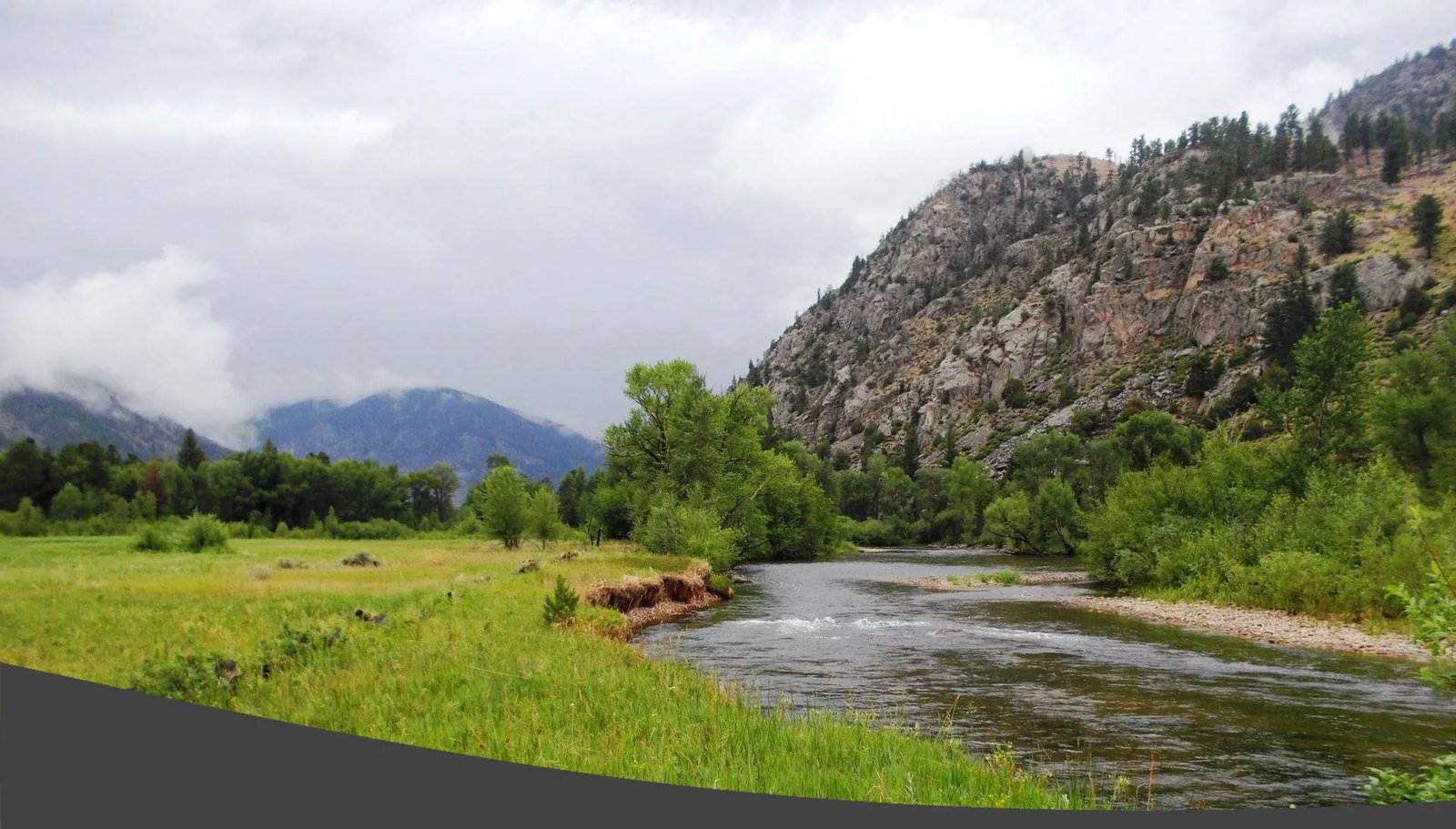 Wide view of a river winding through a green valley with rocky mountains under a cloudy sky