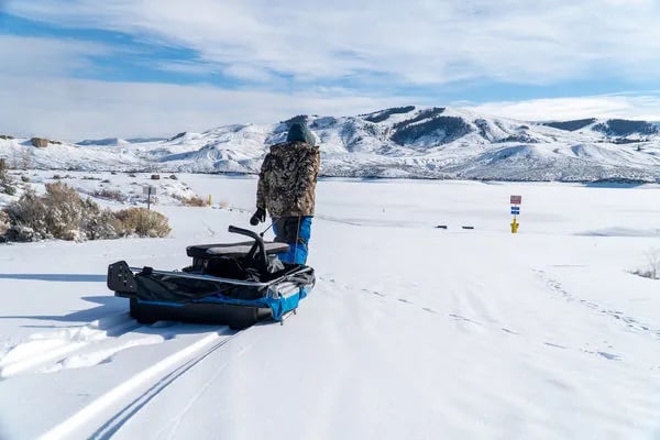 Angler pulling an ice fishing sled across a vast snowy landscape with mountains