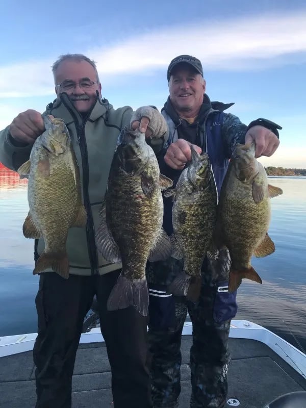 Man on a boat holding two Smallmouth Bass against a calm lake and blue sky.