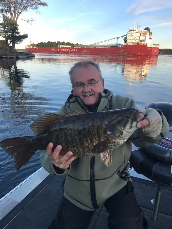 Two men on a boat holding four Smallmouth Bass with a calm lake in the background.