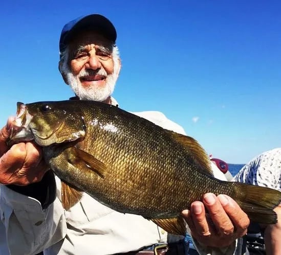 Man with a white beard holding a large Smallmouth Bass against a bright blue sky.