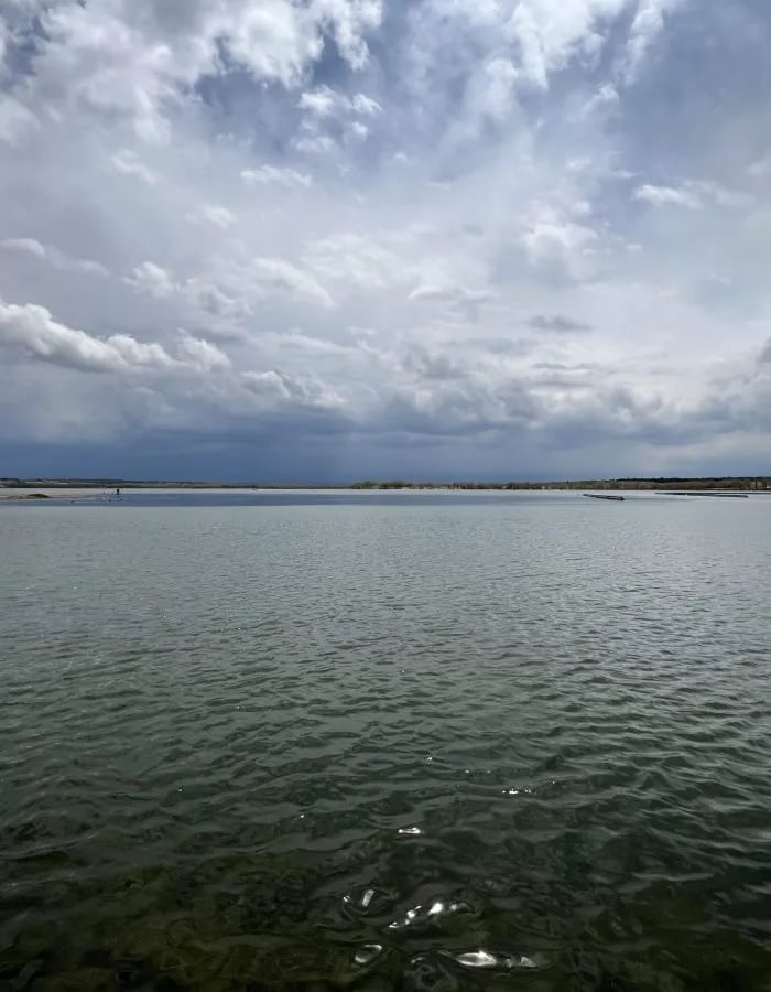 Wide view of a calm lake under a cloudy sky with distant land.
