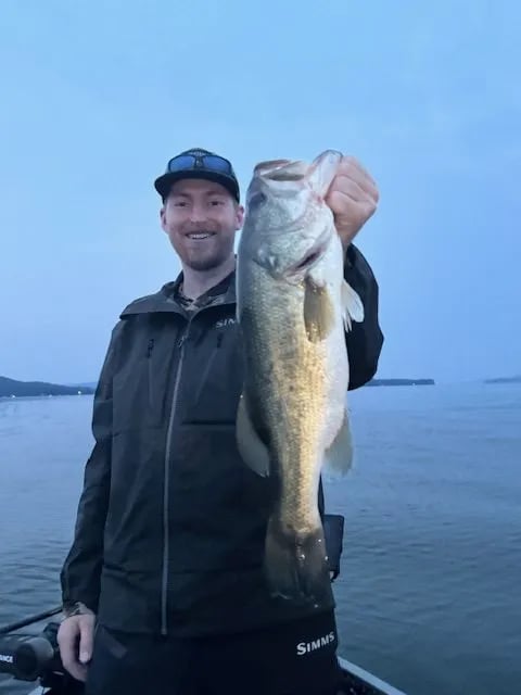 Older man in a white t-shirt holding a large Largemouth Bass on a boat