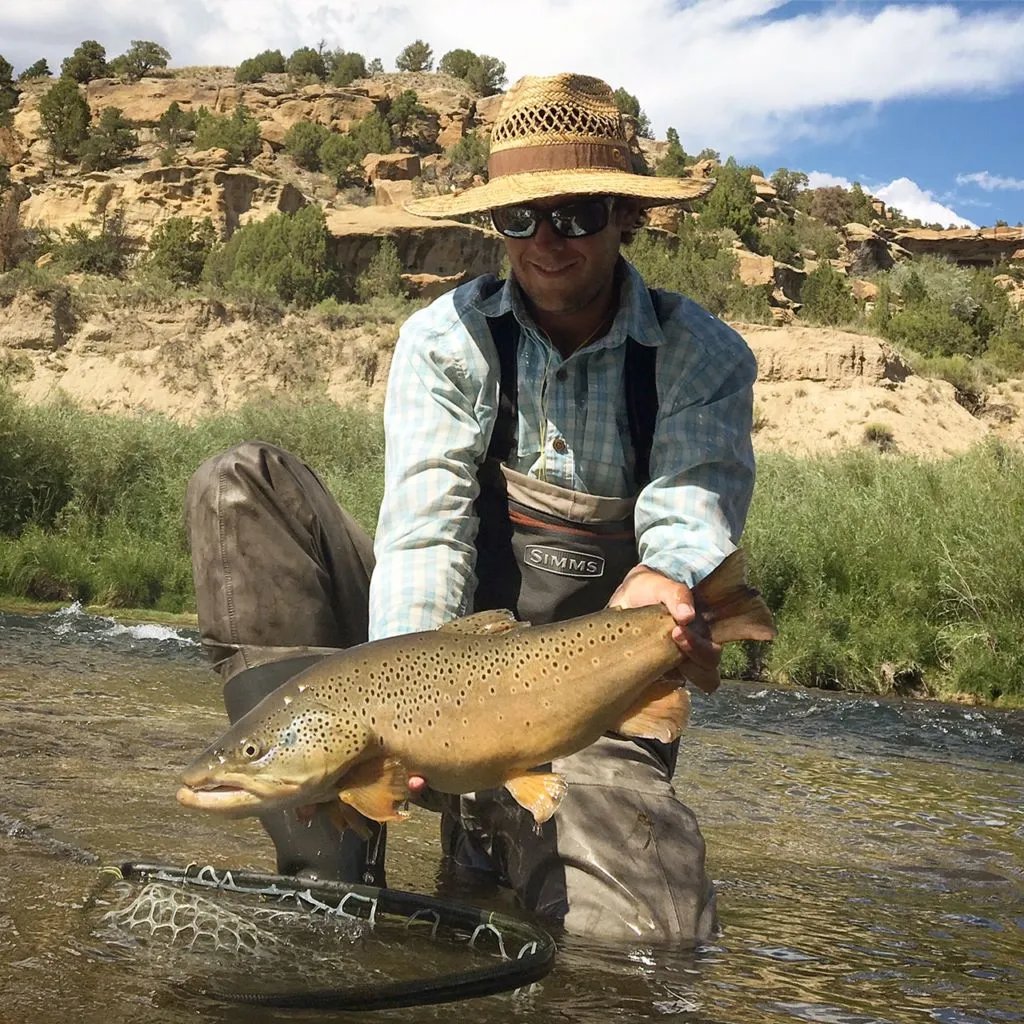 Man in a straw hat holding a large brown trout in a shallow river.