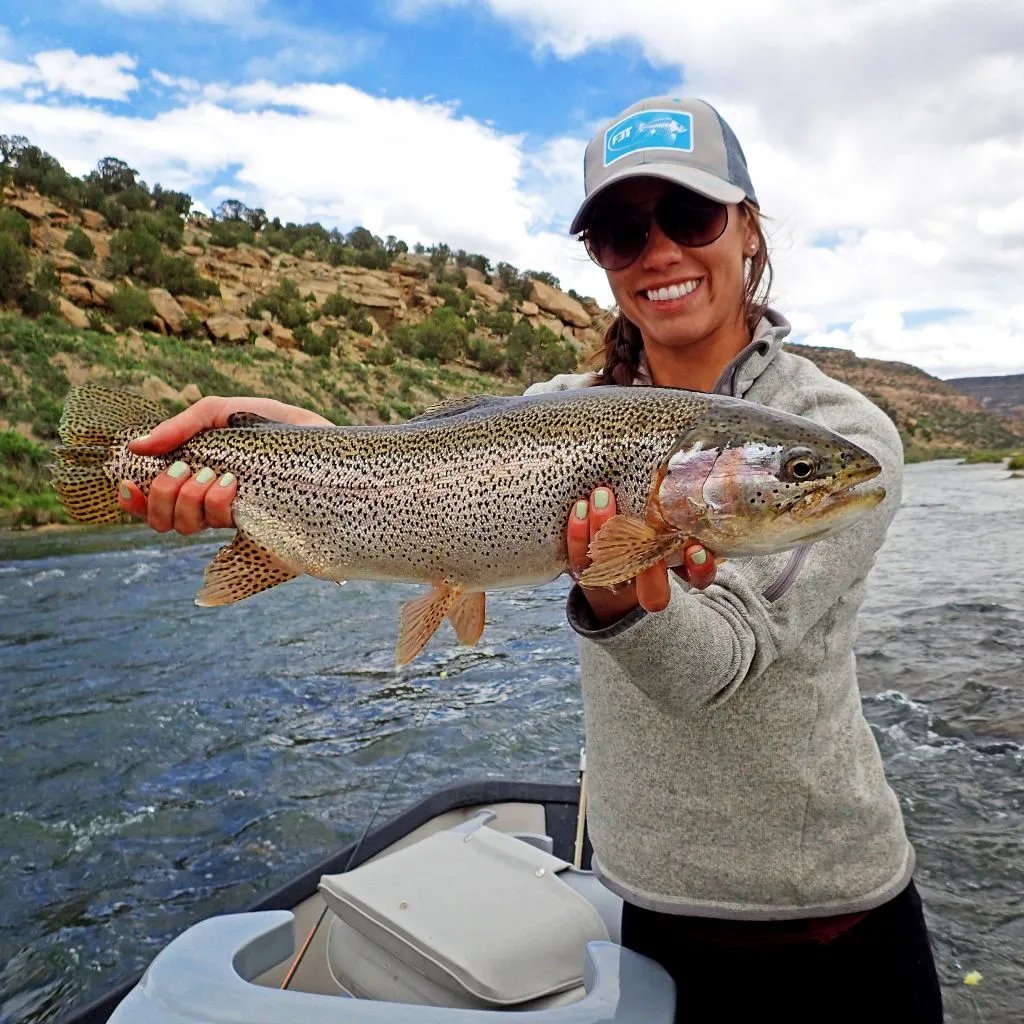 Woman in a baseball cap smiling while holding a large rainbow trout on a boat.