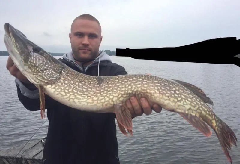 Young man in a bucket hat holding a large Northern Pike with a treeline in the background.