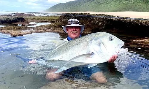 Angler in a hat holding a large trevally in shallow ocean water.