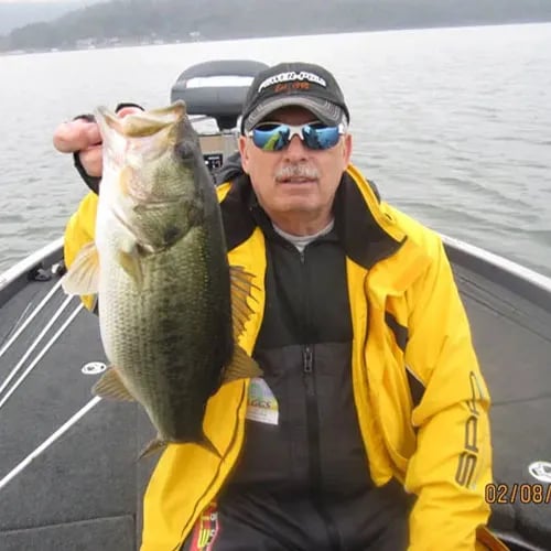 Angler in a light blue shirt holding a large Largemouth Bass on a cloudy lake.