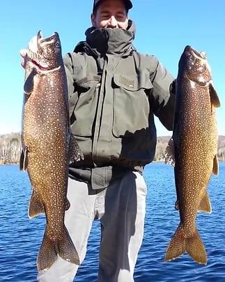 Angler holding two large Lake Trout against a blue sky over water.