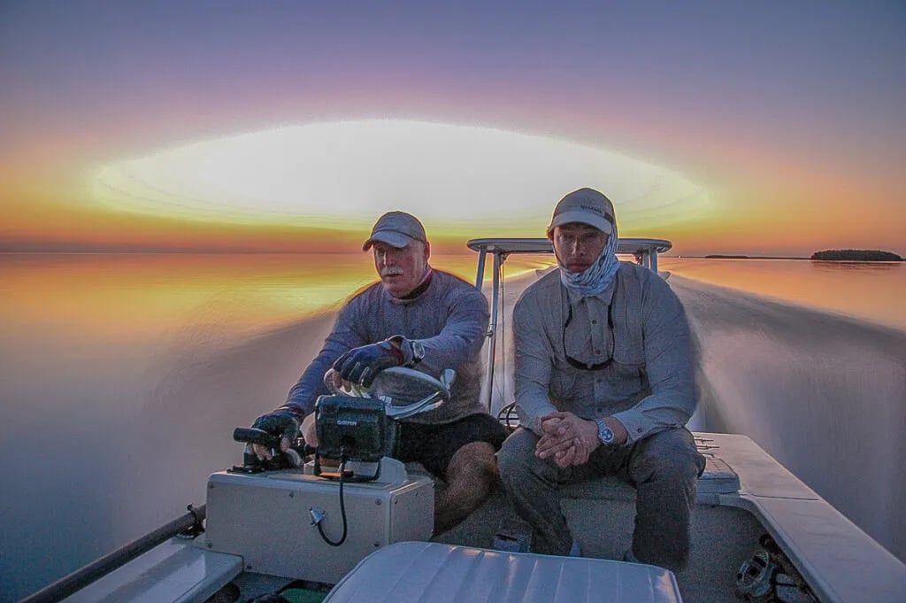 Two anglers on a flats boat driving into a vibrant sunset over calm water.