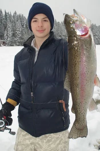 Angler in a blue jacket holding a large rainbow trout in a snowy forest.