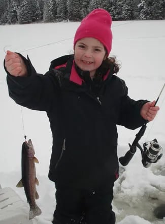Child in a pink hat holding a small trout on an icy lake.