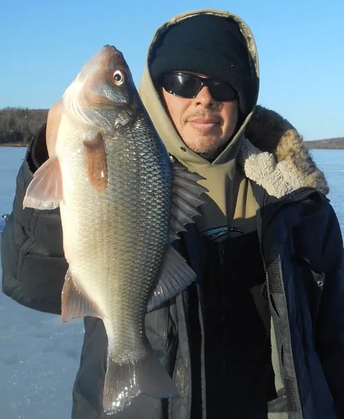 Angler in a winter coat holding a white perch on a frozen lake.