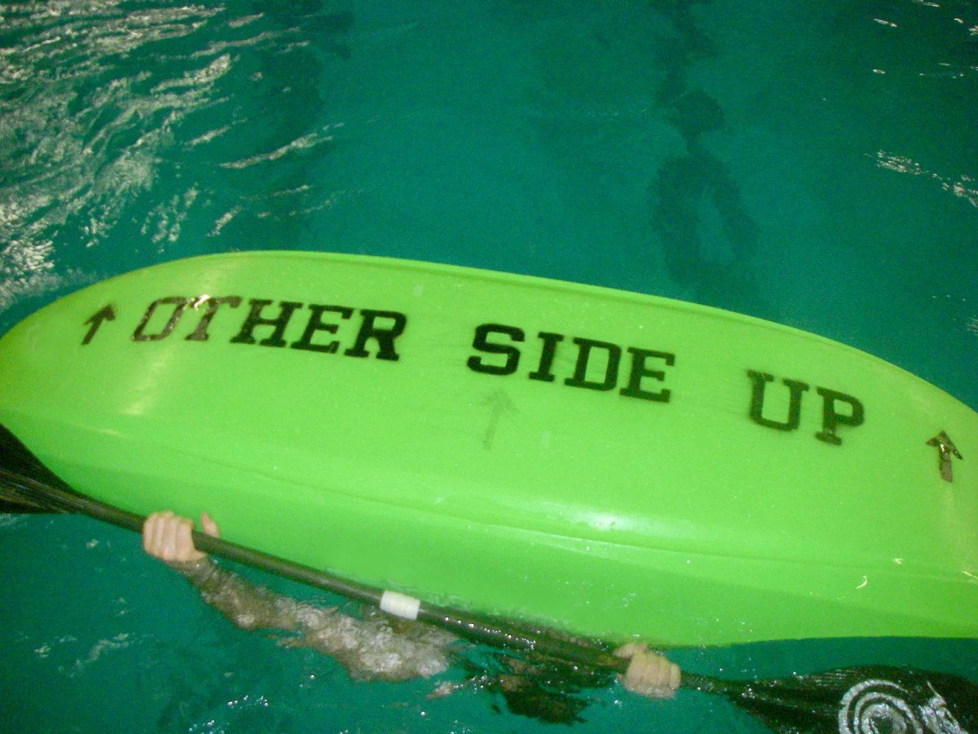 Green kayak upside down in clear water with "OTHER SIDE UP" written on the hull.