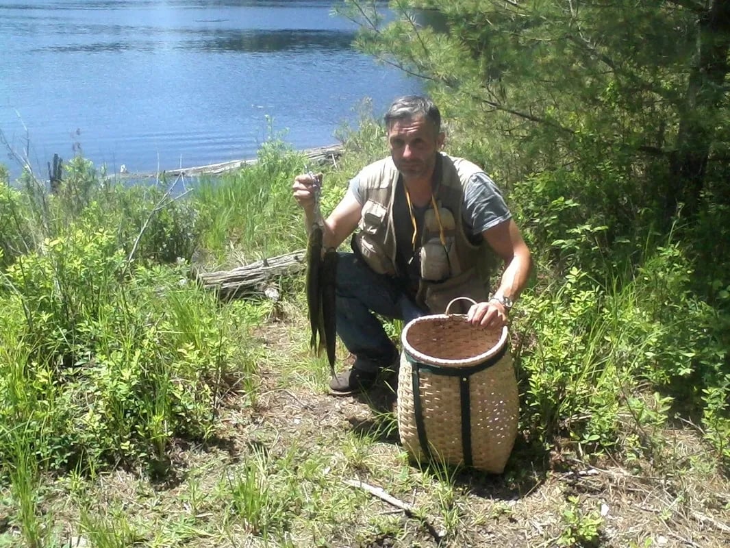 Man holding a Northern Pike next to a woven basket by a lake.
