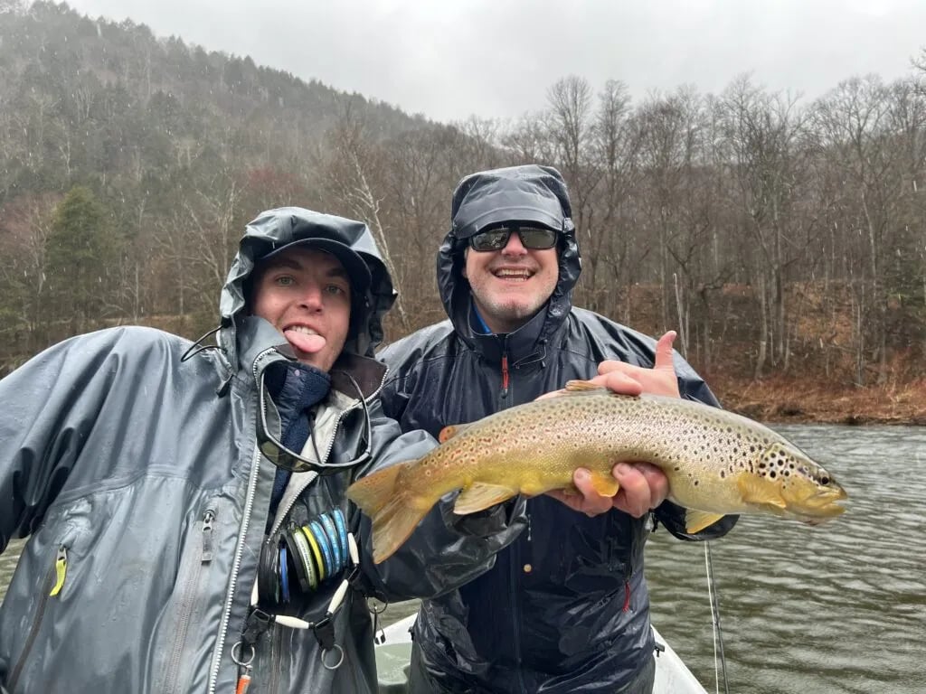 Two anglers in rain gear holding a spotted Brown Trout on a rainy river.