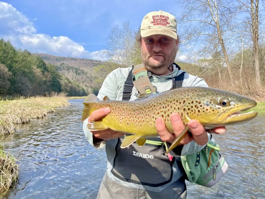 Angler in waders holding a large spotted Brown Trout in a shallow river.