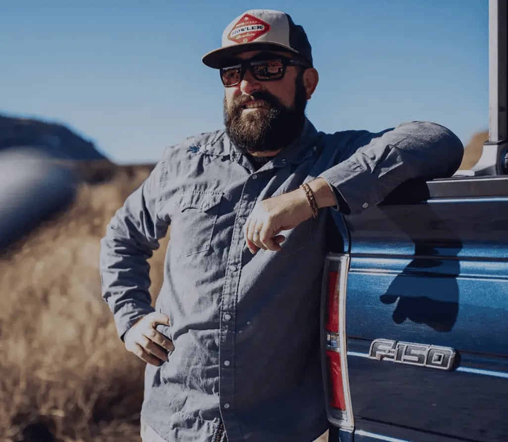 Bearded man in a cap and shirt leaning on a blue truck.