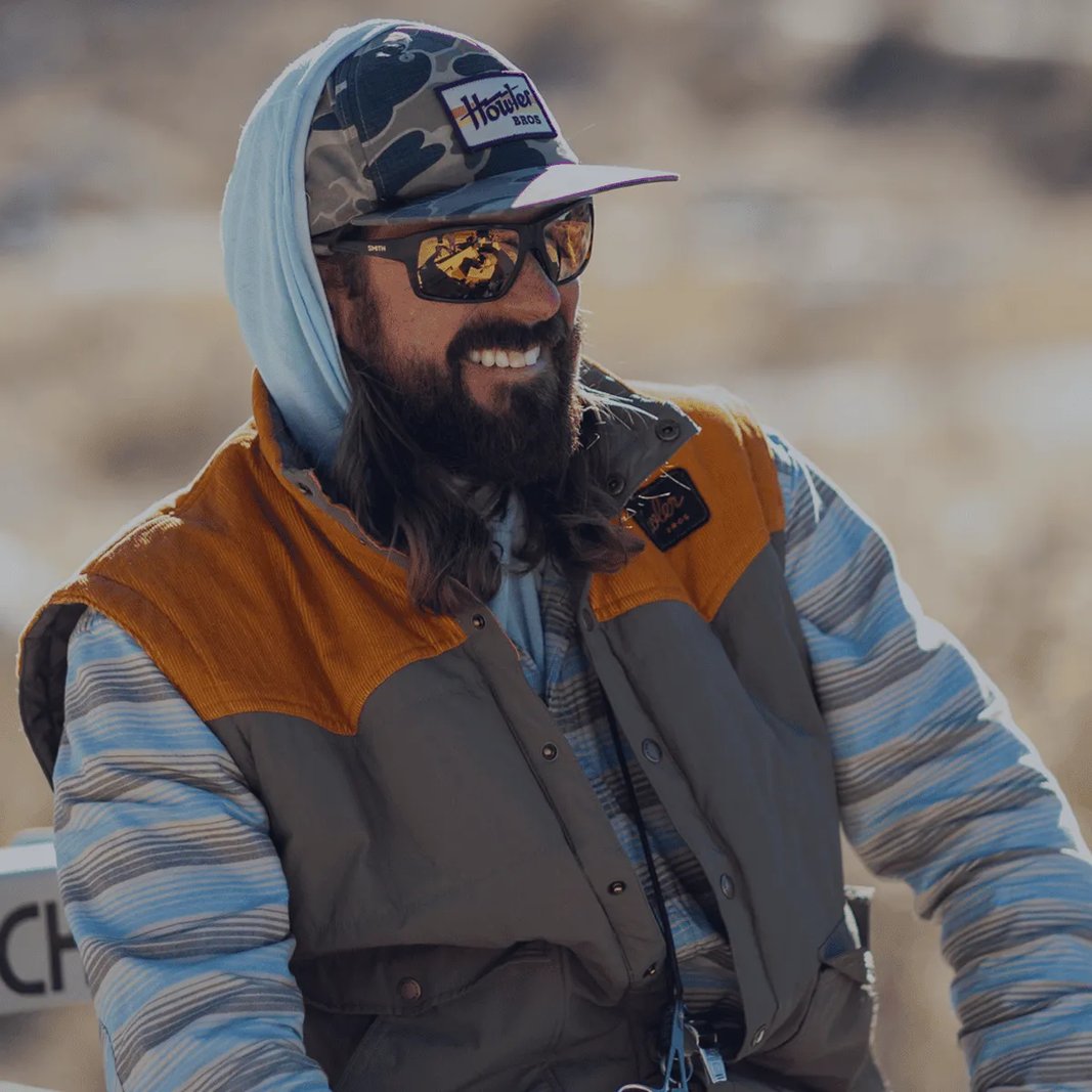 Bearded man in a camo cap and vest smiling outdoors.
