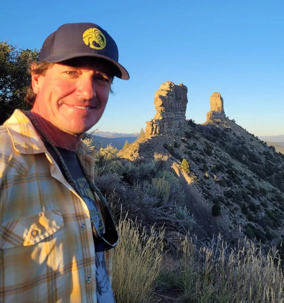 Man in a plaid shirt and cap smiling with rock formations in the background.