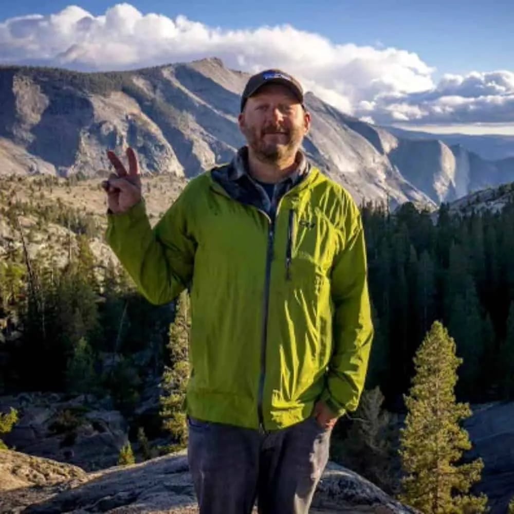 Man in a green jacket giving a peace sign with mountains behind him.