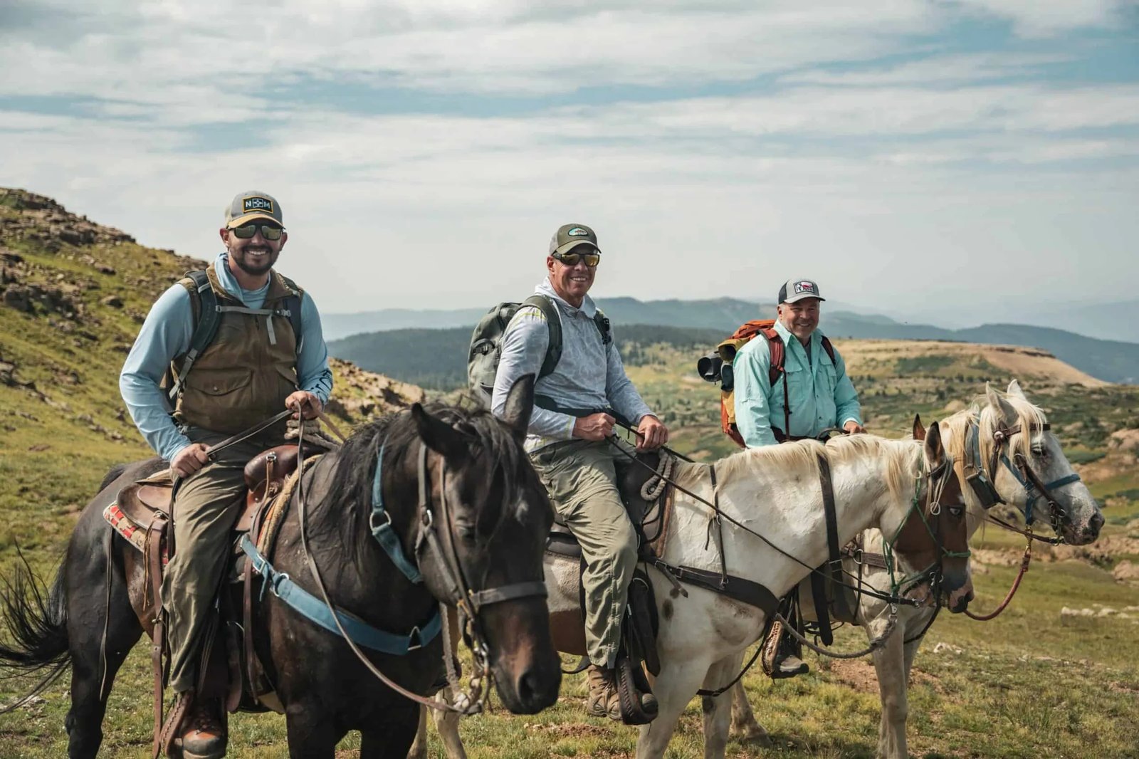 Three people on horseback riding through a grassy mountain landscape.