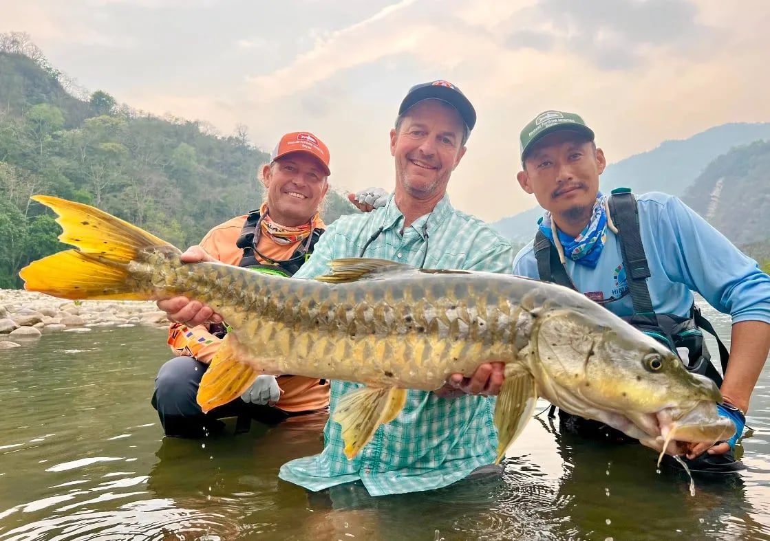 Three anglers in a river holding a large Mahseer with forested mountains in the background.