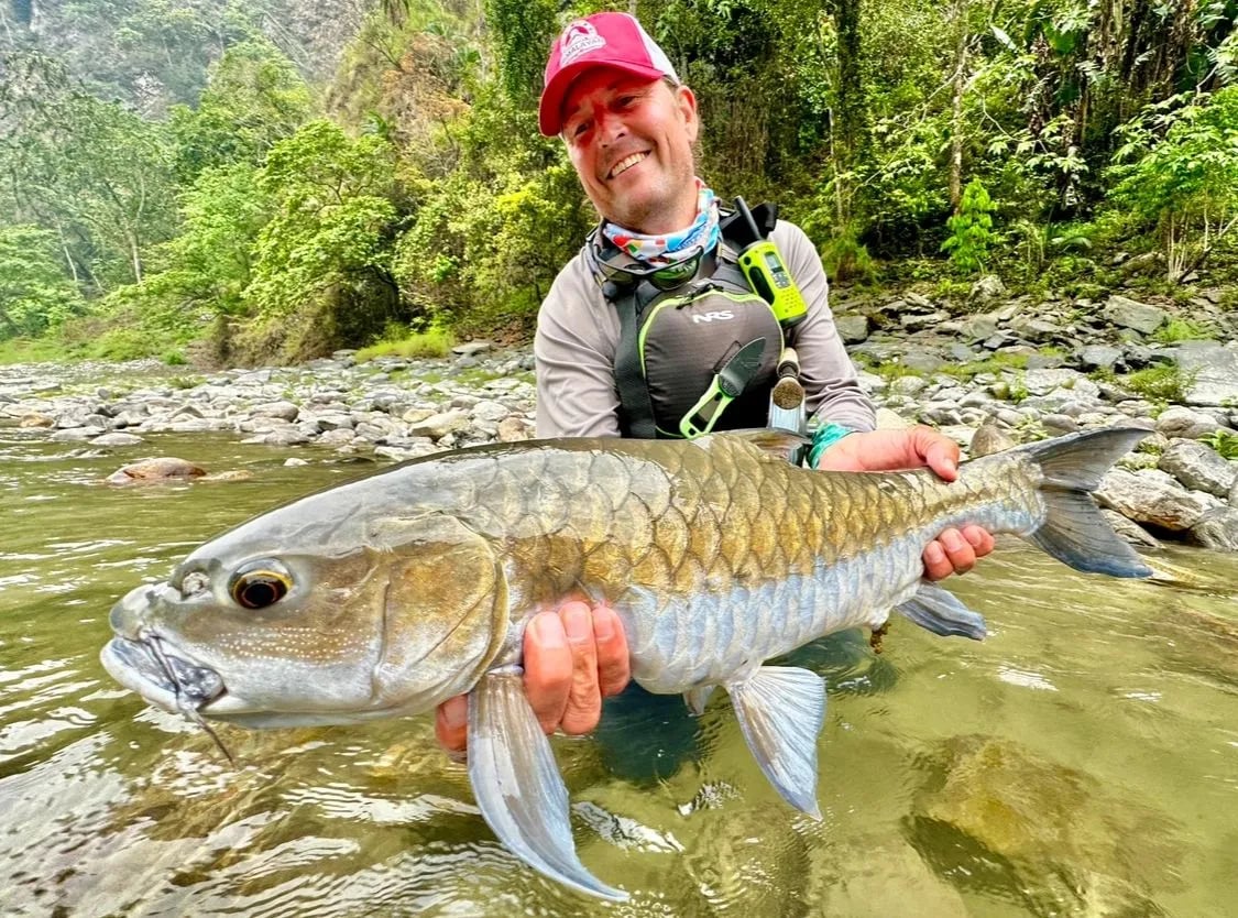 Angler in a river holding a Mahseer with lush green jungle behind.