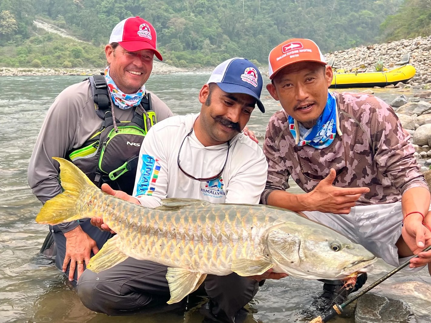Three anglers in a river holding a Mahseer, with a yellow raft on the bank.