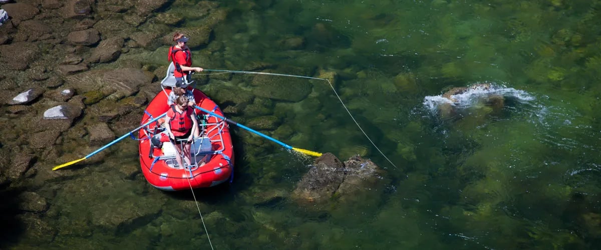 Two anglers fly fishing from a red raft on clear river water, seen from above.