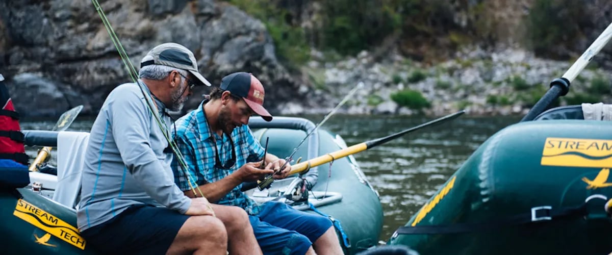 Two men in an inflatable raft, one rigging a fly rod on a river.