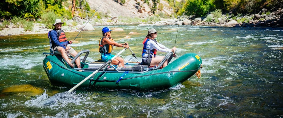 Green inflatable raft and wooden drift boat floating near a rocky bank on a river with rapids.