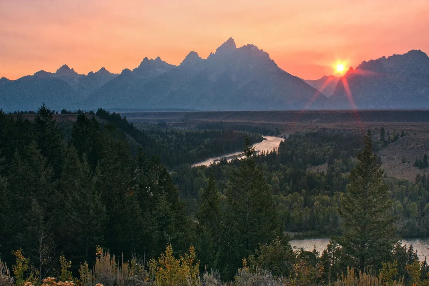 Sunset over a wide river valley with distant mountains and forests.