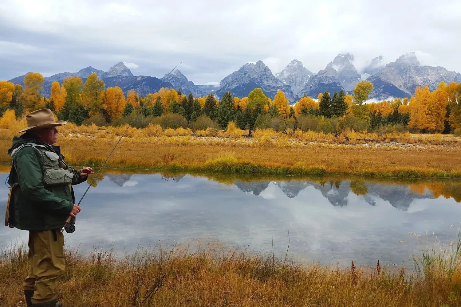 Angler fly fishing in a river with golden autumn trees and snow-capped mountains.