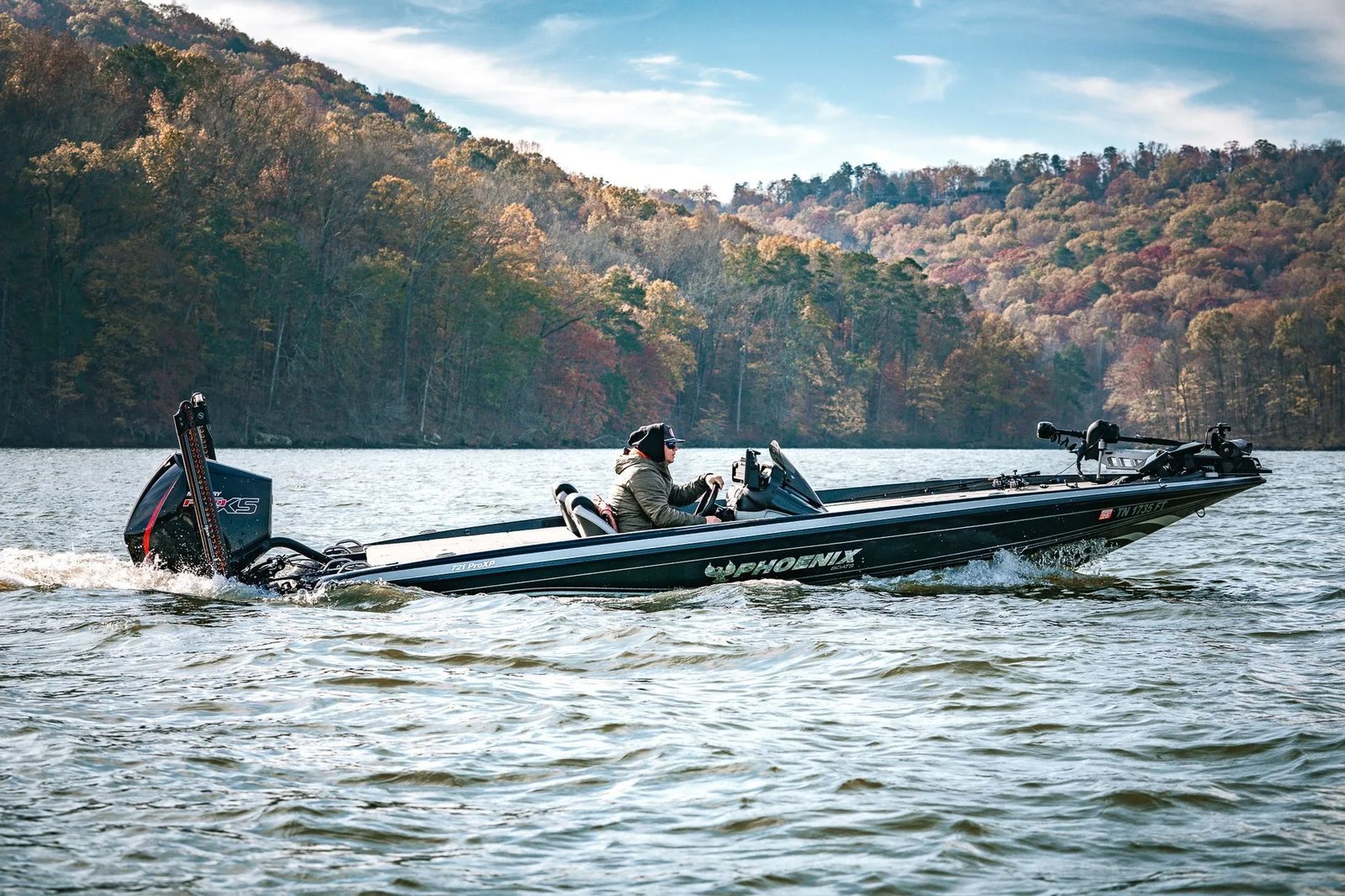 Angler casting from the front of a bass boat on a lake with autumn trees in the background