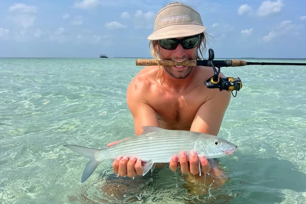Angler holding a bonefish in shallow water with a fly rod in mouth.