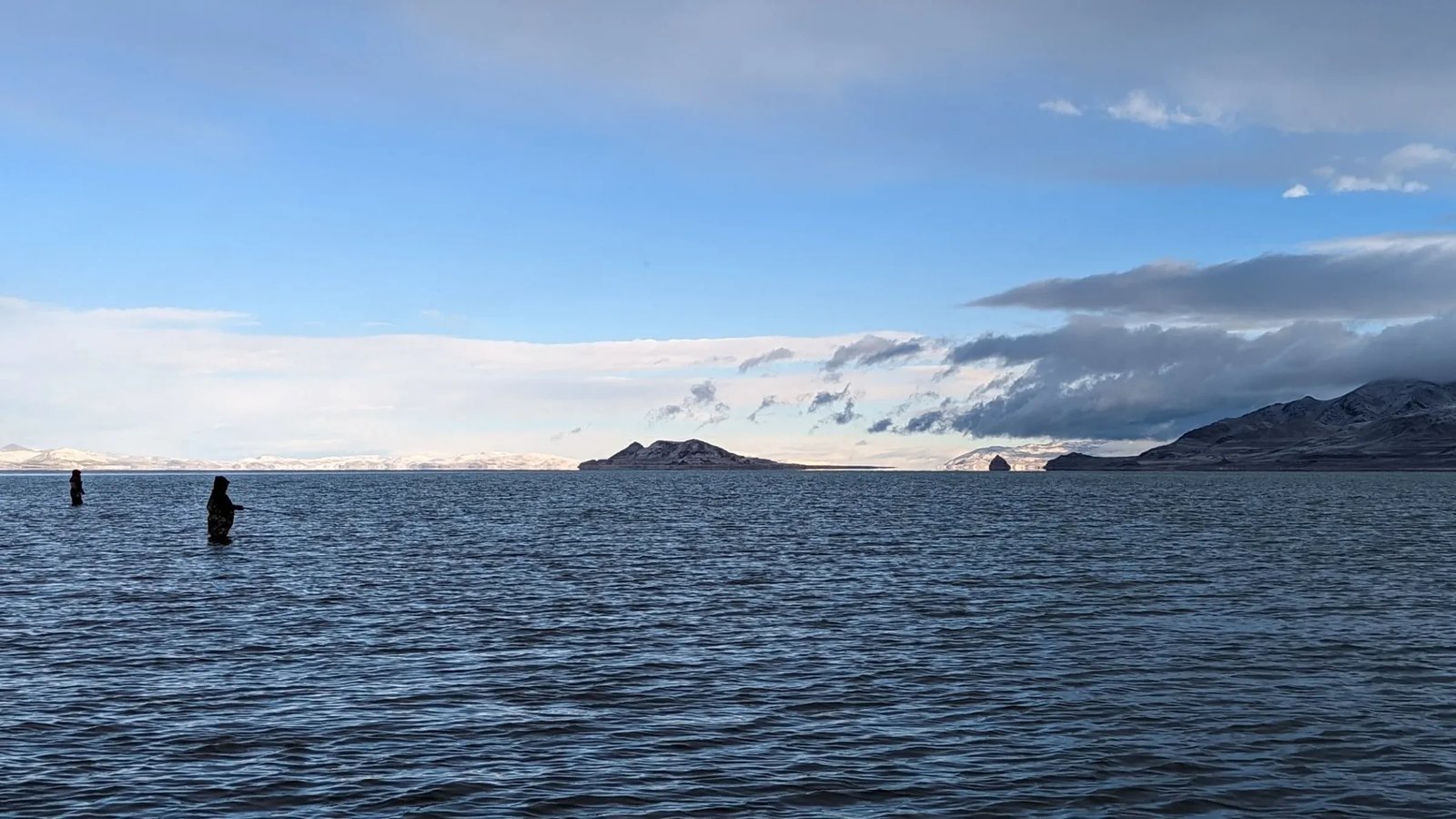 Two anglers wading in a vast lake with distant mountains under a cloudy sky.